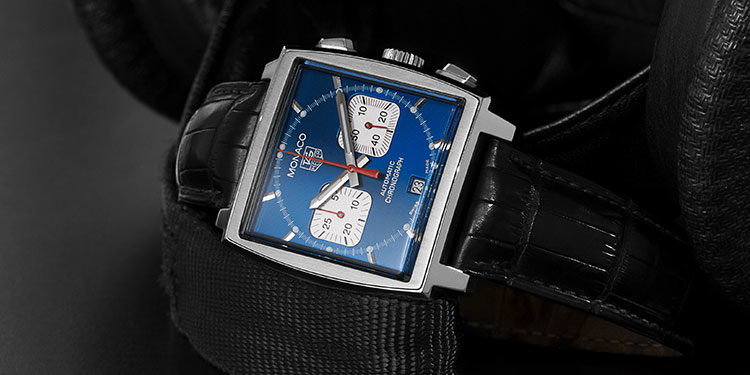 Image: https://dmzr5ikm7nos4.cloudfront.net/content_image/1c37f580-6417-11e8-bb90-596d1c0b64d6/origin.jpg TAG Heuer Monaco mit blauem, rechteckigem Zifferblatt und Lederarmband liegend auf schwarzer spiegelnder Oberfläche
